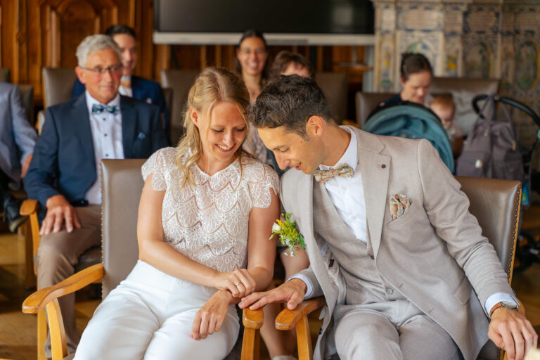 Hochzeitsfotograf Allgäu und Kempten. Braut und Bräutigam sitzen beim Standesamt. Sie schauen beide auf ihren Ringe an den Händen auf der Armlehne zwischen ihnen.