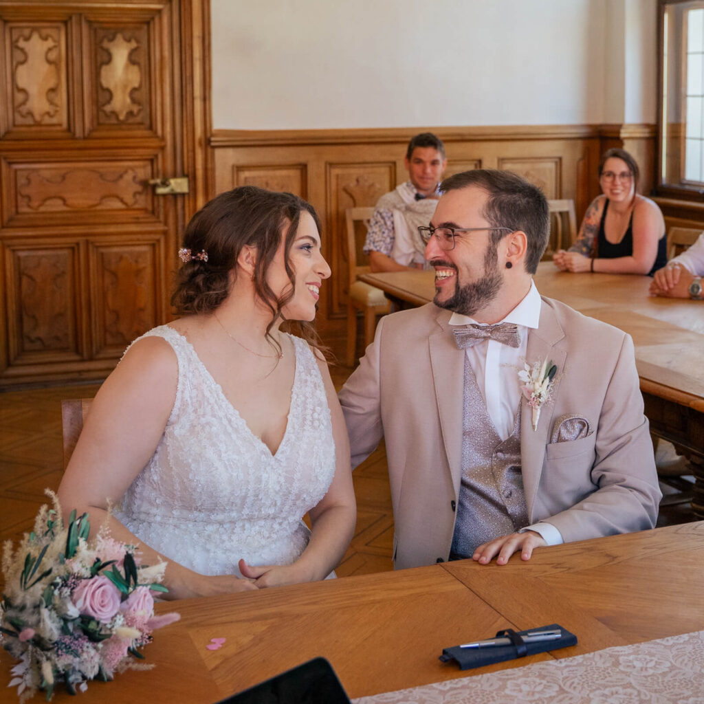 Hochzeitsfotograf Kempten und Allgäu. Ein Brautpaar sitzt beim Standesamt an einem Tisch. Die Braut trägt ein weißes Brautkleid. Das Paar schaut sich in die Augen. Der Bräutigam hat seinen Arm um die Braut gelegt. Die Gäste schauen im Hintergrund zu.