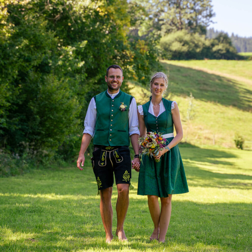 Hochzeitsfotograf Kempten und Allgäu. Brautpaar in Tracht beim Brautpaarshooting. Das Brautpaar läuft Hand in Hand auf einer grünen Wiese am Waldrand auf den Betrachter zu. Beide lächeln. Die Braut hält ihren Brautstrauß in den Händen