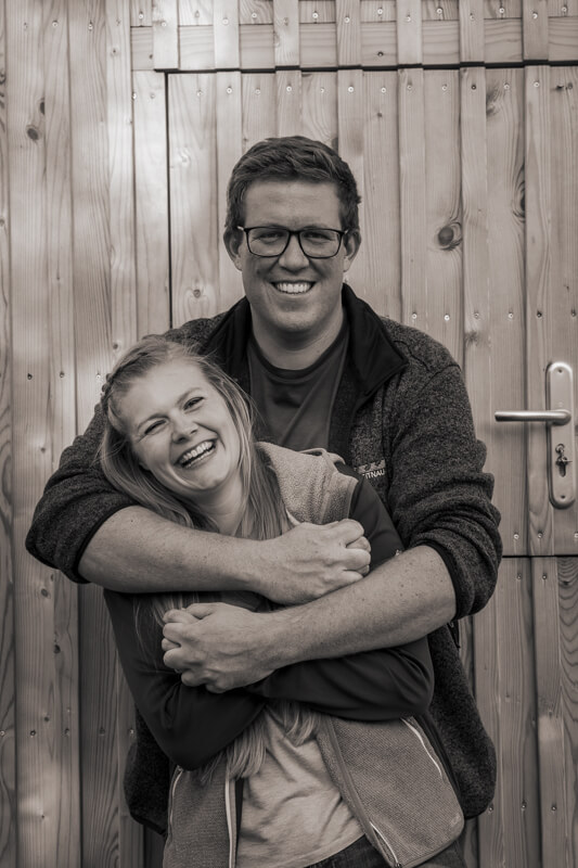 Hochzeitsfotograf Kempten und Allgäu. Ein Paar steht hintereinander vor einen Holzwand. Der Mann steht hinter der Frau und hat seine Arme um sie geschlungen. Beide lachen.