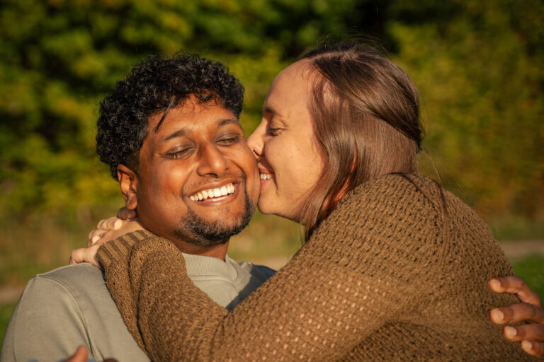 Paarshooting vom Fotograf im Allgäu. Ein Paar genießt die Zweisamkeit in der Spätsommerabendsonne. Die Frau hat die Arme um den Hals des Mannes gelegt und gibt ihm mit geschlossenen Augen einen Kuss auf die Wange. Der Mann lächelt dabei mit geschlossenen Augen und hält sein Gesicht in die Sonne.