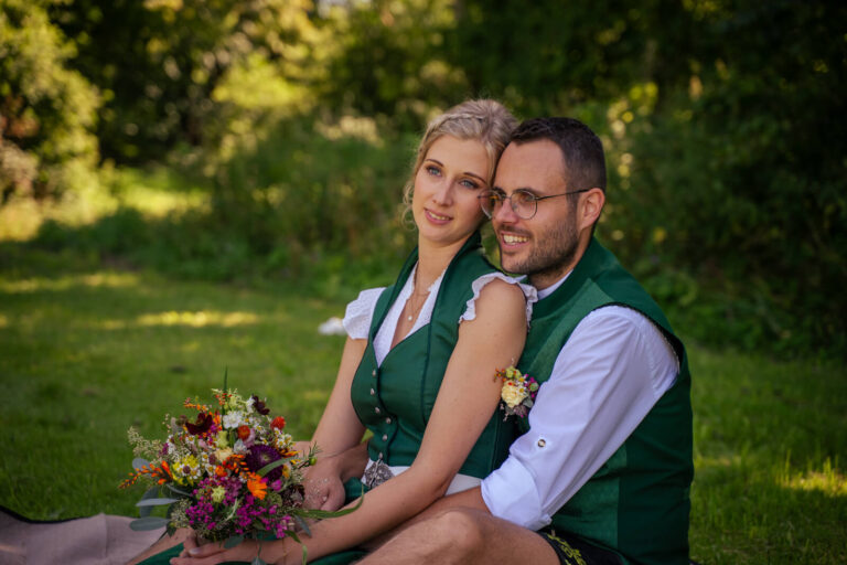Hochzeitsfotograf Allgäu und Kempten. Ein Brautpaar in Tracht sitzt hintereinander auf einer Wiese. Der Mann sitzt hinter der Frau. Die Frau hält ihren Brautstrauß. Gemeinsam schauen sie in die Ferne.