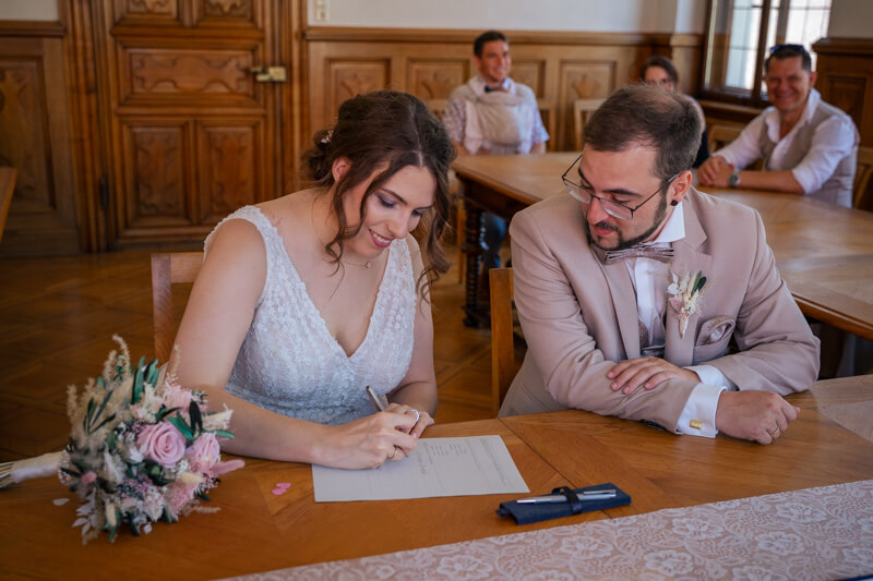 Hochzeitsfotograf Allgäu und Kempten. Ein Brautpaar unterschreibt im Standesamt die Dokumente. Die Frau hält einen Stift in der Hand und sieht auf ein Blatt Papier. Der Mann schaut ihr dabei zu.
