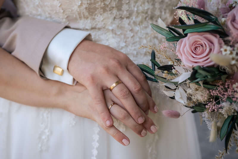 Hochzeitsfotograf Allgäu und Kempten. Eheringe an den Händen des Brautpaares. Die Hand des Mannes hält dabei die Hand der Frau. Im Hintergrund sind das Brautkleid und der Brautstrauß.