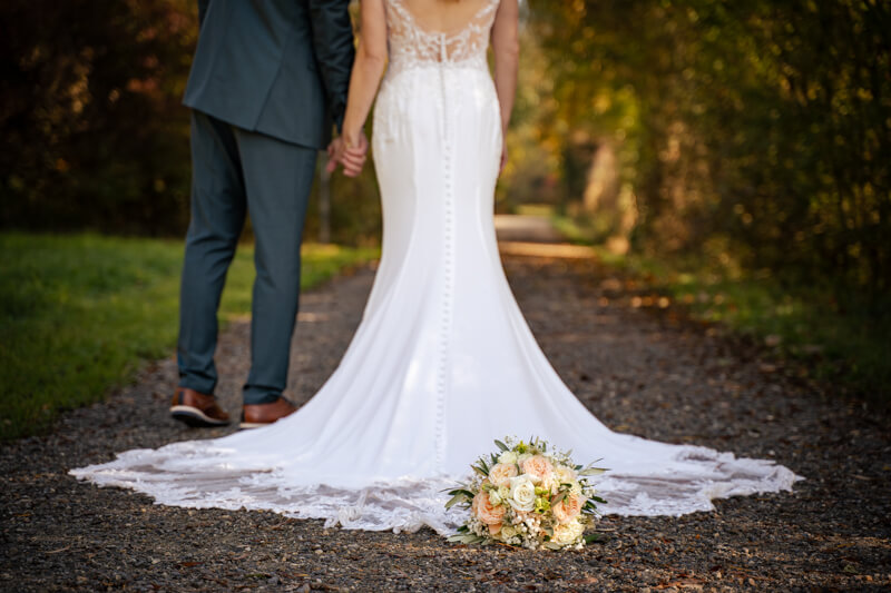 Hochzeitsfotograf Allgäu und Kempten. Ein ausgebreitetes Brautkleid auf einem Kiesweg. Der Brautstrauß liegt am Ende der Schleppe auf dem Kies. Der Bräutigam steht neben der Braut. Von beiden sind nur die Beine zu sehen.