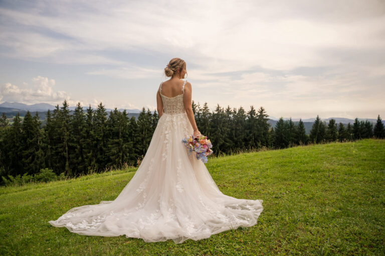 Hochzeitsfotograf Allgäu und Kempten. Die Braut steht auf einer Wiese. Dahinter ist Wald. Die Braut trägt ein weißes langes Brautkleid mit einer langen Schleppe. Die Schleppe liegt ausgebreitet am Boden. Die Braut lässt ihren Arm mit dem Brautstrauß locker hängen. Sie schaut in die Ferne.