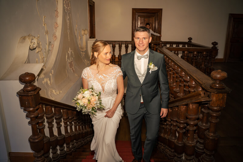 Hochzeitsfotograf Allgäu und Kempten. Ein Brautpaar geht Seite an Seite die Treppe zum Trauzimmer im Standesamt hoch. Der Mann schaut zum Betrachter, die Braut seiht seitlich nach unten. Sie hält ihren Brautstrauß in der Hand.